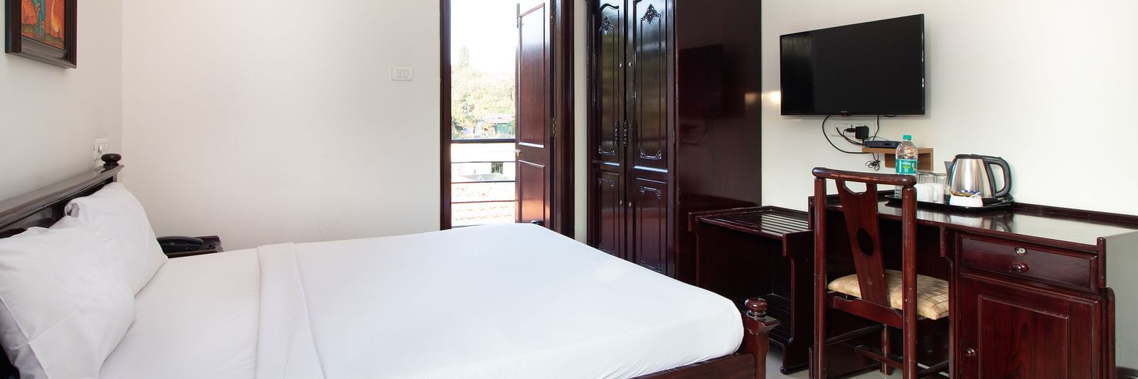 A bright room with a double bed with white linens, complemented by dark wooden furniture and a doorway leading to the balcony at Sanctum Suites Indiranagar, Bangalore.