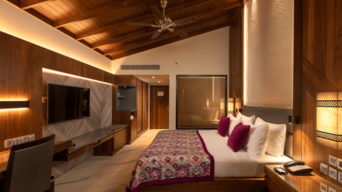 A king-size bed in front of TV inside Premium Club Room at Kenilworth Resort & Spa, Goa, with side lamps, side tables, a ceiling fan, and a glass wall of the bathroom. The resort is ideal for South Goa long-term stays with modern amenities and premium faci