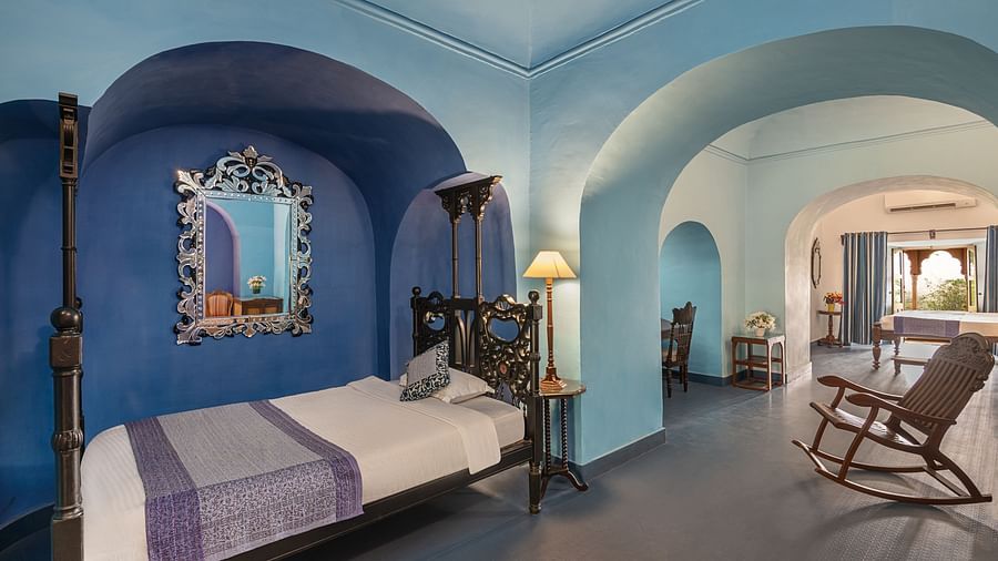 Room with bed, patterned runner, mirror in arched alcove, lamp, rocking chair, and view into another room with bed, curtains, and arched doorway - Tijara Fort-Palace - 19th Century, Alwar.