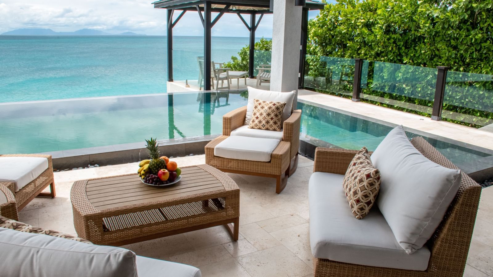 A luxurious outdoor patio with a seating area and an infinity pool overlooking a turquoise ocean and lush green trees under a bright sky.