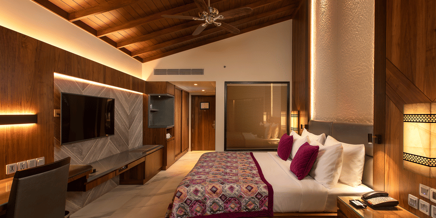 A king-size bed in front of TV inside Premium Club Room at Kenilworth Resort & Spa, Goa, with side lamps, side tables, a ceiling fan, and a glass wall of the bathroom. The resort is ideal for South Goa long-term stays with modern amenities and premium faci