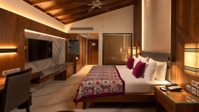 A king-size bed in front of TV inside Premium Club Room at Kenilworth Resort & Spa, Goa, with side lamps, side tables, a ceiling fan, and a glass wall of the bathroom. The resort is ideal for South Goa long-term stays with modern amenities and premium faci