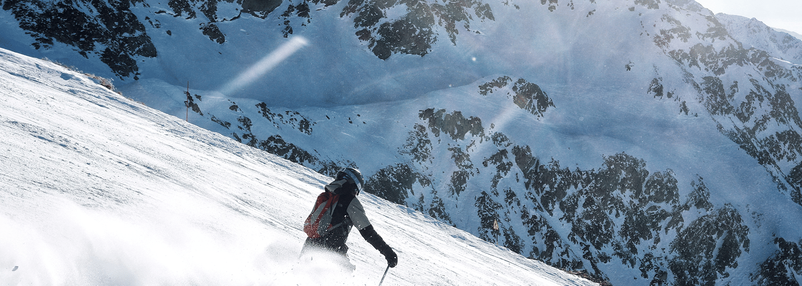 A skier descending a steep, snow-covered mountain slope on a sunny day, kicking up powdery snow against a rocky alpine background.