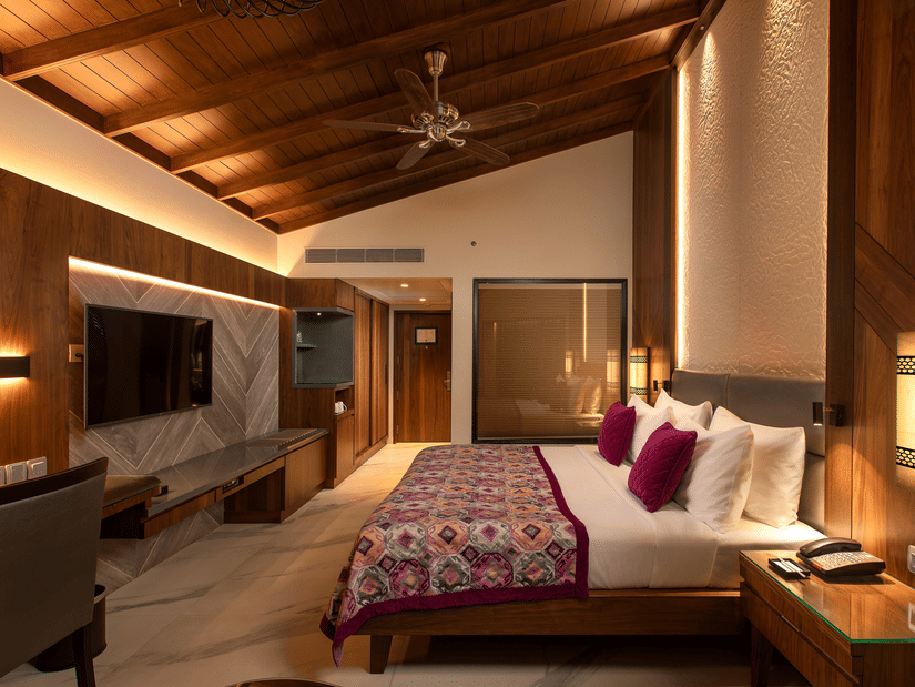 A king-size bed in front of TV inside Premium Club Room at Kenilworth Resort & Spa, Goa, with side lamps, side tables, a ceiling fan, and a glass wall of the bathroom. The resort is ideal for South Goa long-term stays with modern amenities and premium faci