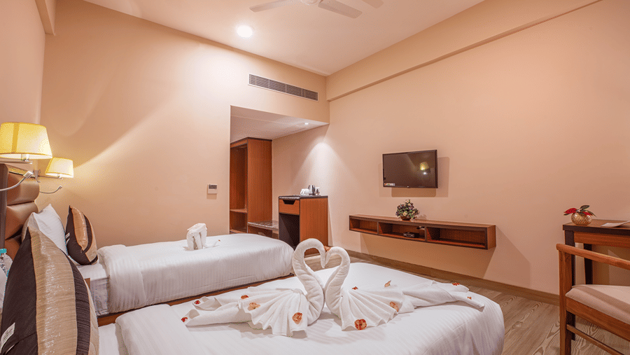 A modern bedroom at The Lalita Grand Mathura - Vrindavan featuring twin beds, swan towel art on white bedding, a wall-mounted TV, and ambient lighting.