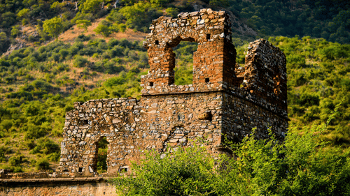 Ruined stone structures with arched windows situated on a grassy slope with dense green foliage in the background.