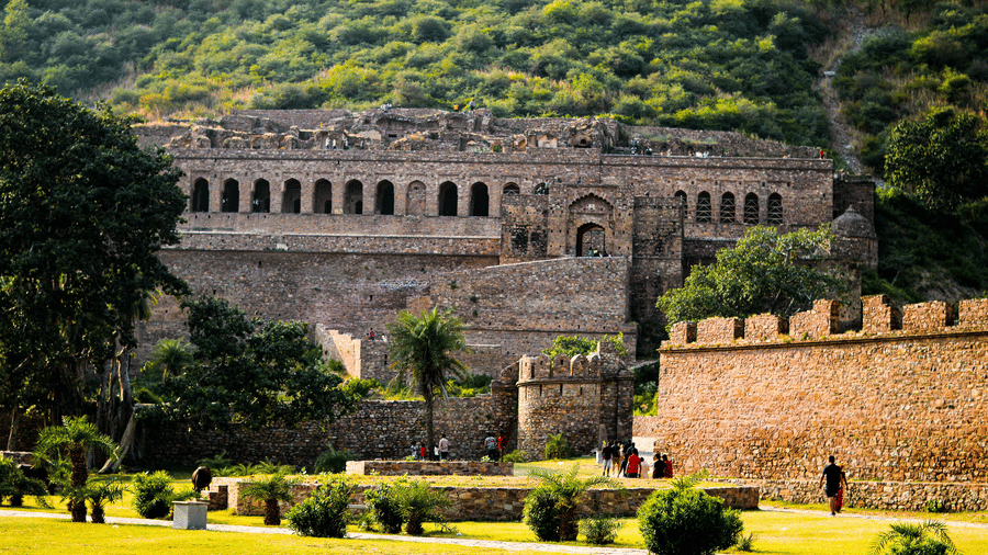 A distant view of Bhangarh fort, featuring a multi-storey fort built into a hillside with arched openings and a green lawn in the foreground.