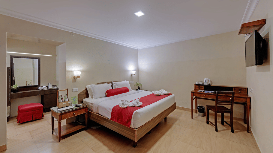 A spacious bedroom at Holiday Home Resort, Kodaikanal, featuring a neatly-made bed, a workspace with a table and a chair, a wall-mounted television, and a wooden dressing table. 