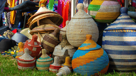 Colourful handwoven coiled baskets in tall round shapes with red, blue, green and yellow patterns arranged on grass at an outdoor market.