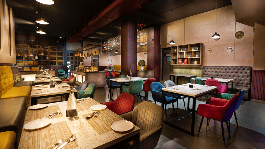 A vibrant restaurant interior with rows of wooden tables, colourful velvet armchairs, and atmospheric lighting from low-hanging pendants at Apartel by Aarin, Oragadam.