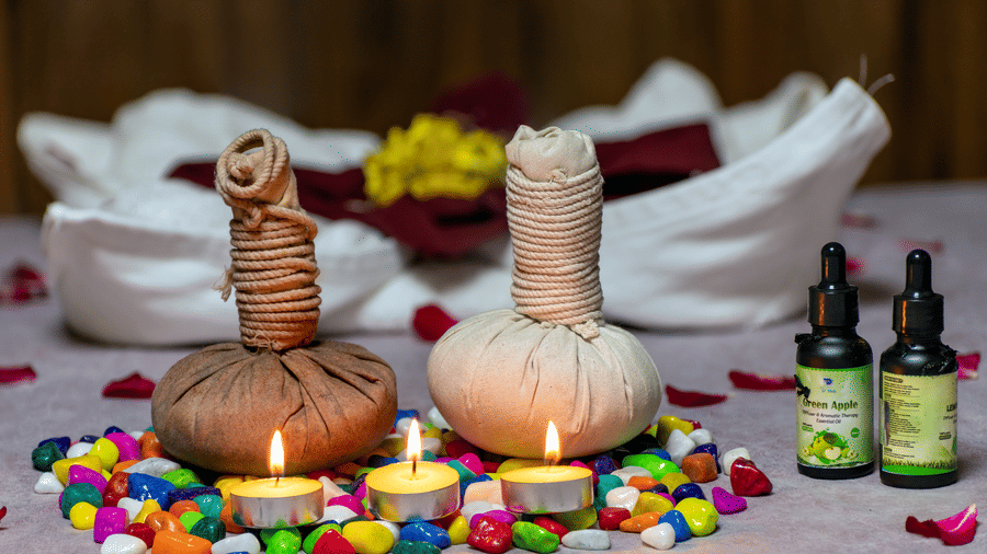 A spa setting with two herbal compress balls, lit candles, essential oil bottles, and rose petals