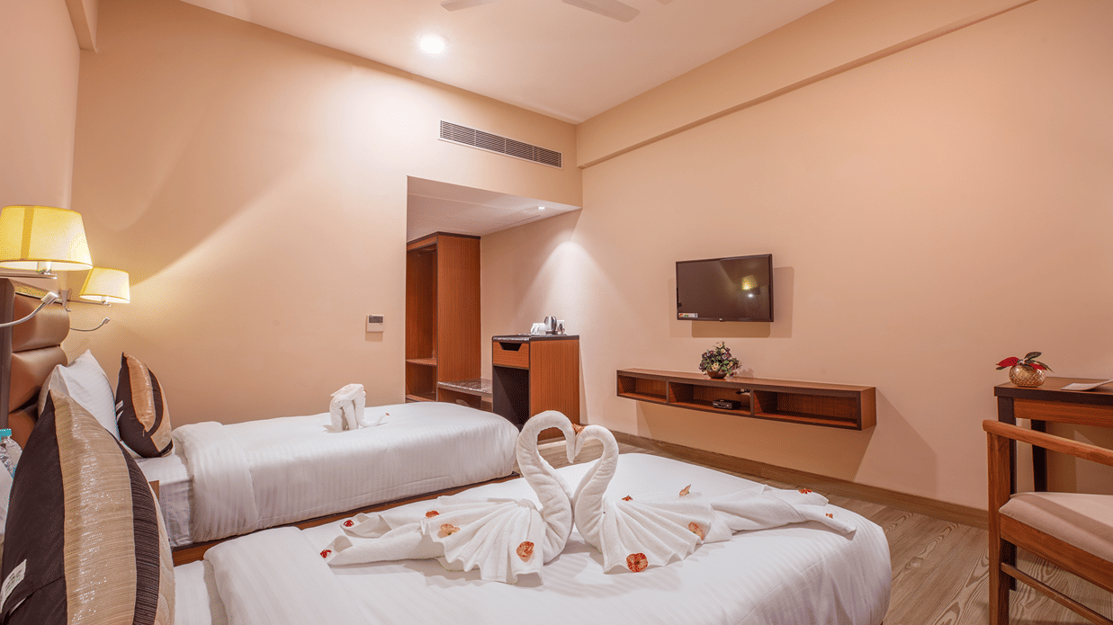 A modern bedroom at The Lalita Grand Mathura - Vrindavan featuring twin beds, swan towel art on white bedding, a wall-mounted TV, and ambient lighting.