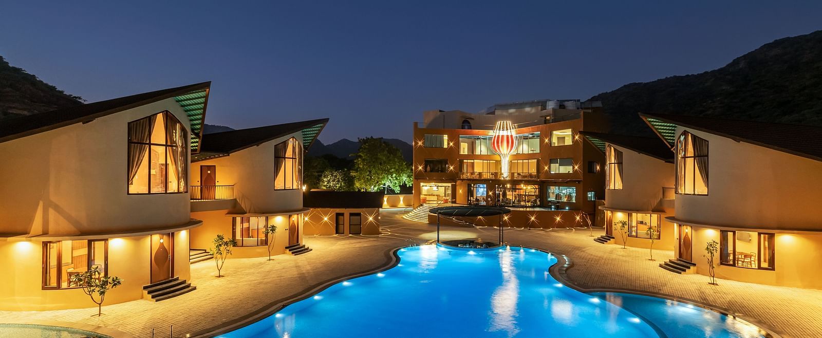 The Qualia Resort Club & Brewery, Udaipur at night with illuminated buildings, a brightly lit swimming pool, and landscaping.