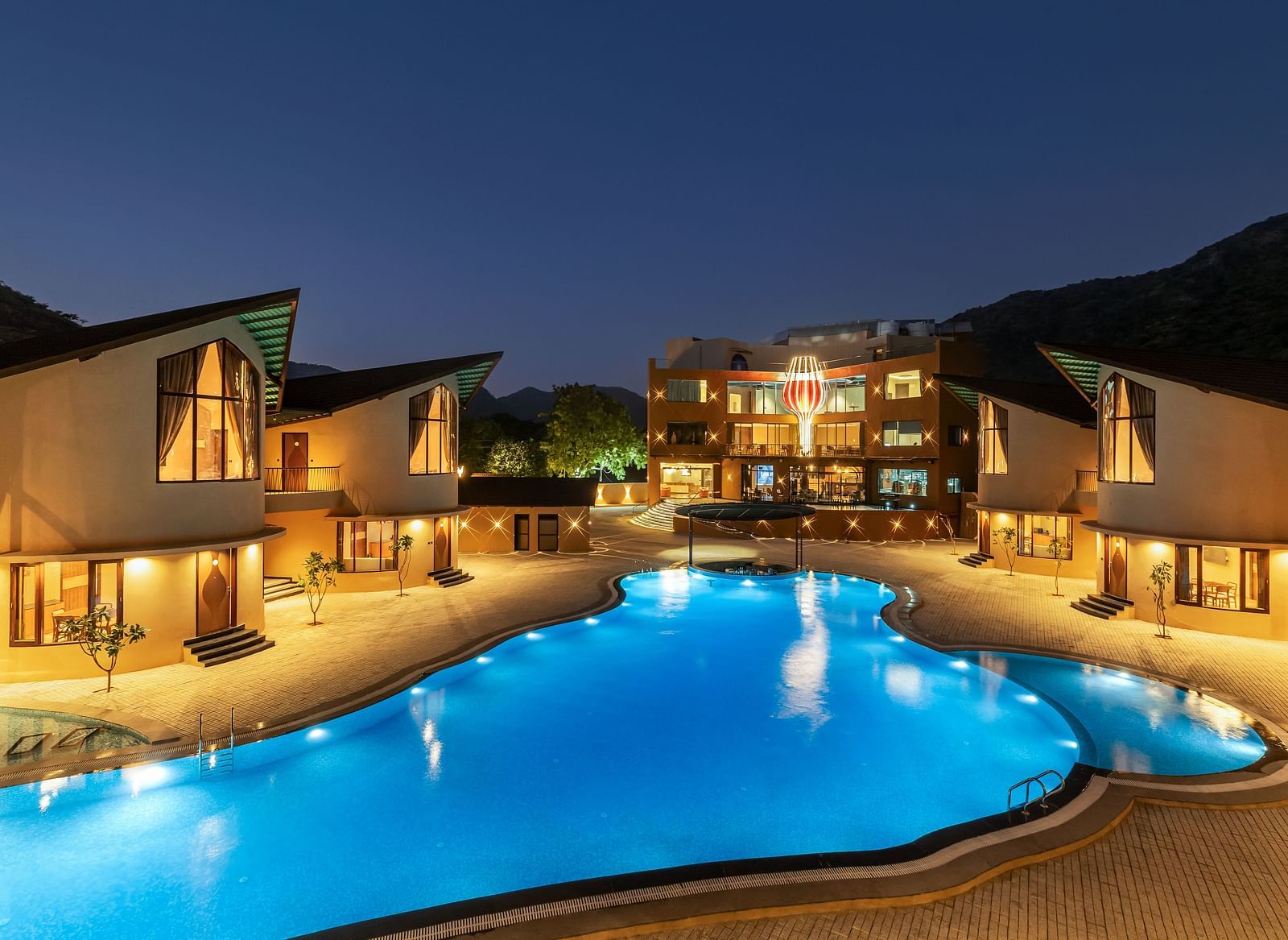 The Qualia Resort Club & Brewery, Udaipur at night with illuminated buildings, a brightly lit swimming pool, and landscaping.