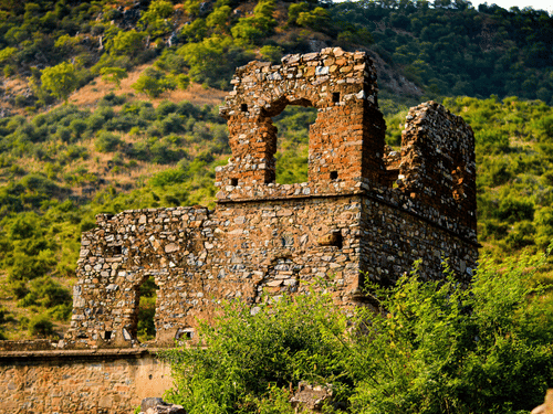 Ruined stone structures with arched windows situated on a grassy slope with dense green foliage in the background.