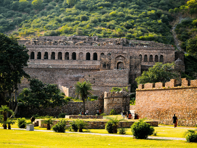 A far-out view of Bhangarh Fort, Rajasthan, with a manicured garden having shrubs and trees and a green mountain behind it.