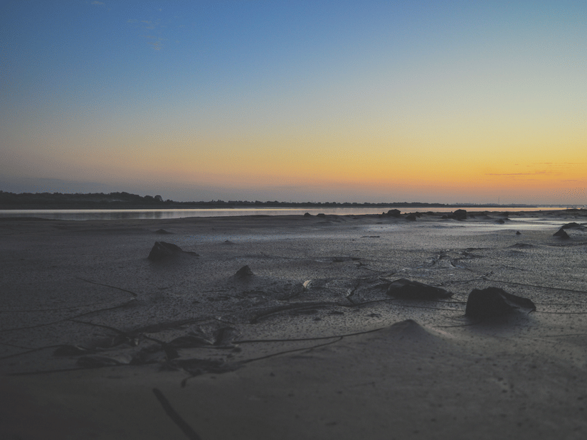 Scenic sunset view over a calm river with rocky shoreline and vibrant evening colours across the horizon.