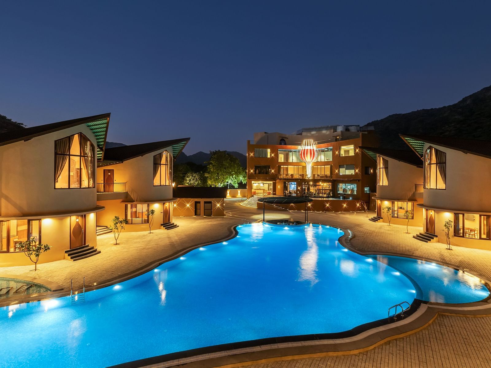 The Qualia Resort Club & Brewery, Udaipur at night with illuminated buildings, a brightly lit swimming pool, and landscaping.