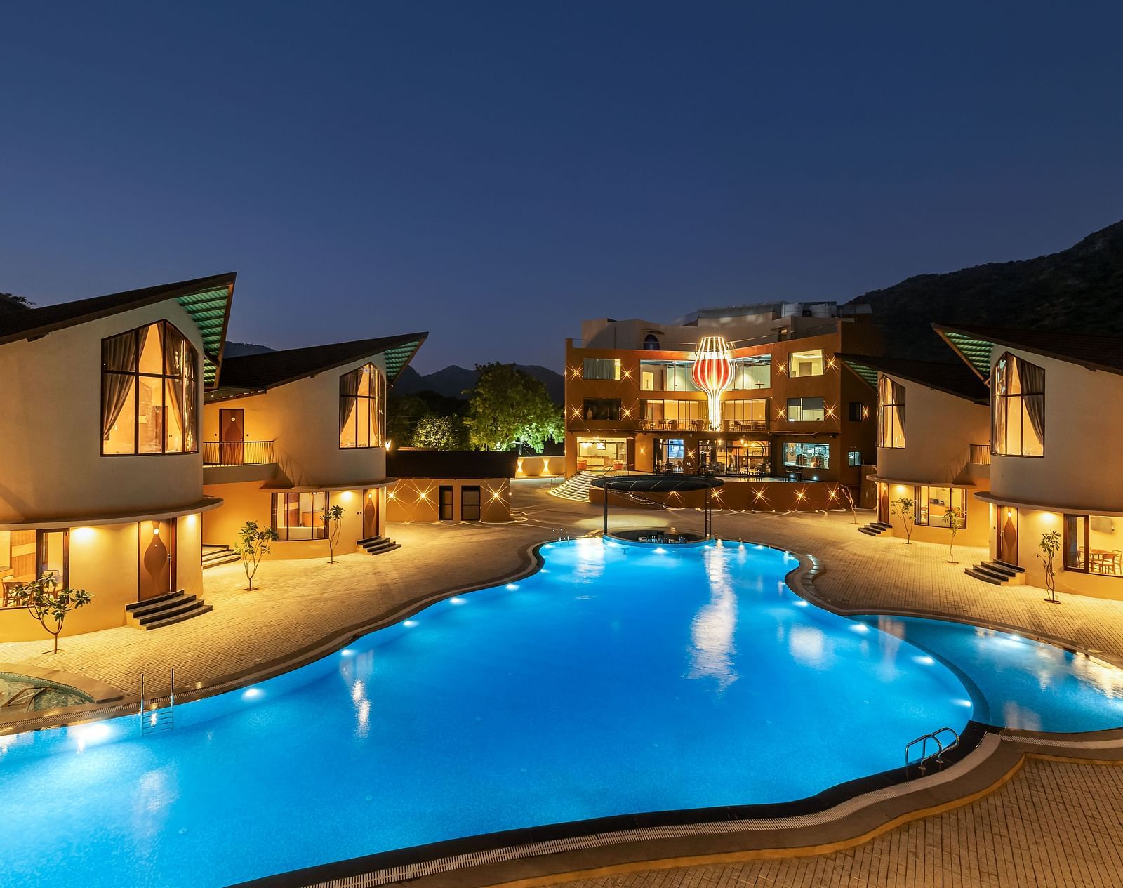The Qualia Resort Club & Brewery, Udaipur at night with illuminated buildings, a brightly lit swimming pool, and landscaping.