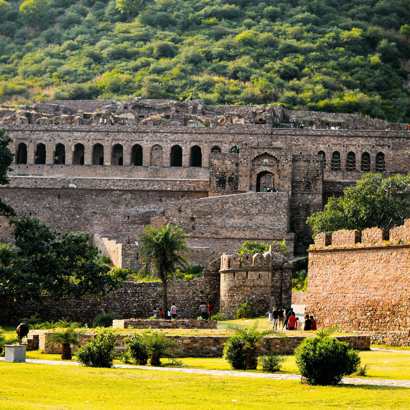 An impressive historic stone fortress stands against a lush green hillside, bordered by a manicured lawn.