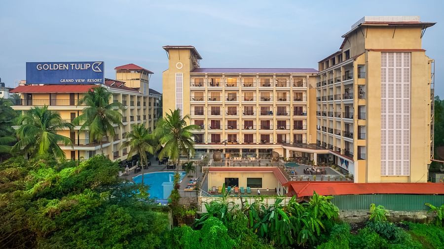 an aerial front angle shot of a hotel in candolim, goa, featuring towering buildings with baclonies, a signage with hotel name, surrounded by lush greenerytowering hotel buildings featuring balconies, a signage with hotel name, and surrounded by lush green