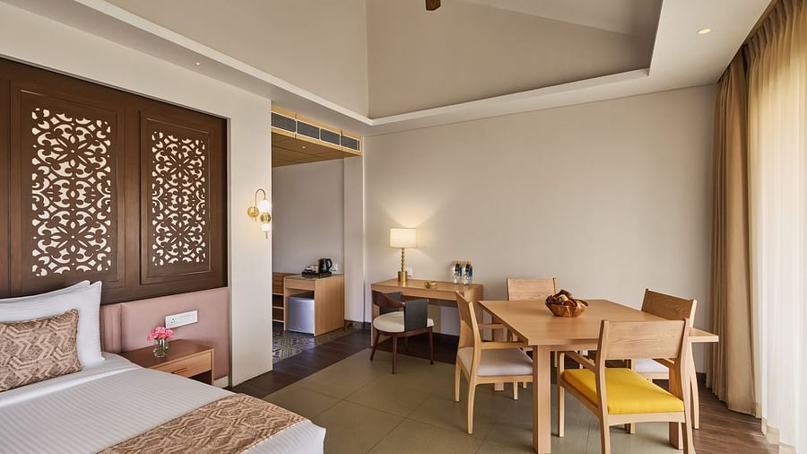Modern hotel room with a cosy bed featuring a decorative wooden headboard, a wooden dining table with chairs, ceiling fan, work desk with a lamp, and floor-to-ceiling curtains allowing natural light - Voyage The Solitaire Bliss.