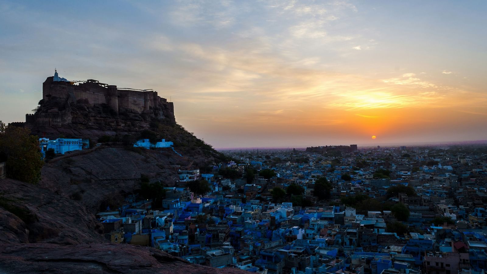 an aerial view of the city of Jodhpur filled with blue buildings with the sun setting in the background