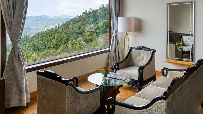  A cosy seating area  at Digantaa Resort in Mukteshwar featuring armchairs and a small table by a large window offering a scenic view of rolling green hills
