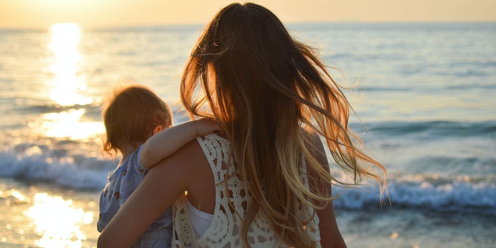 A mother holding her child while standing at the beach, enjoying the serene sunset over the ocean.