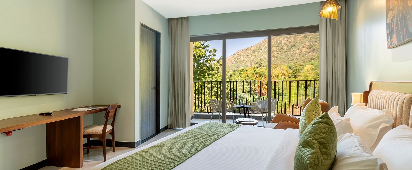 Club Suite with Valley View Sitout showcasing a king size bed with green and white linens, a work desk, and a balcony with a scenic view at The Qualia Resort Club & Brewery, Udaipur.