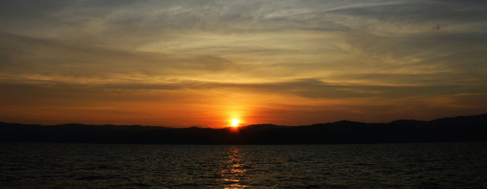 A tranquil sunset over water with the sun dipping behind a mountain range, under a vast, cloud-streaked sky.