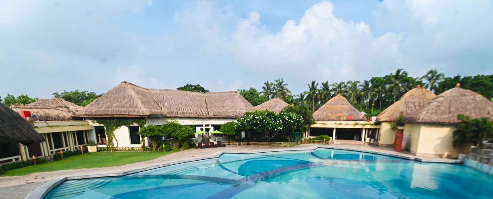 A daylight view of the common swimming pool at Vedic Village Spa Resort, featuring buildings with tatched roofs in the background.