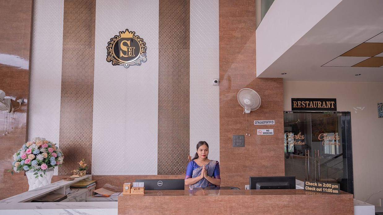 The reception area of Clarks Collection Somnath with marble desk, flower arrangement, staff member, and signage on the wall.