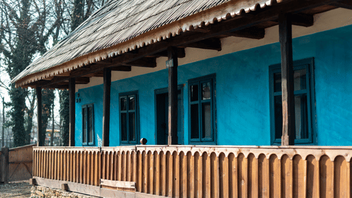 Exterior of a single-story house with blue walls, wooden railing, and a sloped roof, set among trees