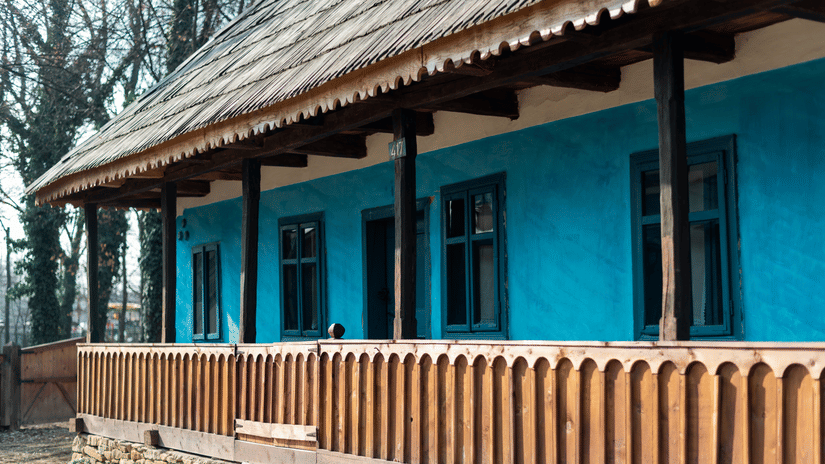 Exterior of a single-story house with blue walls, wooden railing, and a sloped roof, set among trees