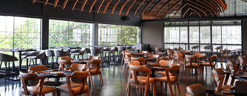 Contemporary restaurant dining room at Hotel Daspalla, Hyderabad featuring curved illuminated wooden ceiling, wooden chairs, dark tables, and floor-to-ceiling windows with garden views.