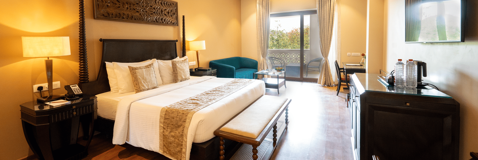 Premium Room at Essentia Luxury Resort & Spa, Udaipur showing a plush bed and an ottoman at its foot facing the TV over a table on the wall with a seating space by the door to the balcony