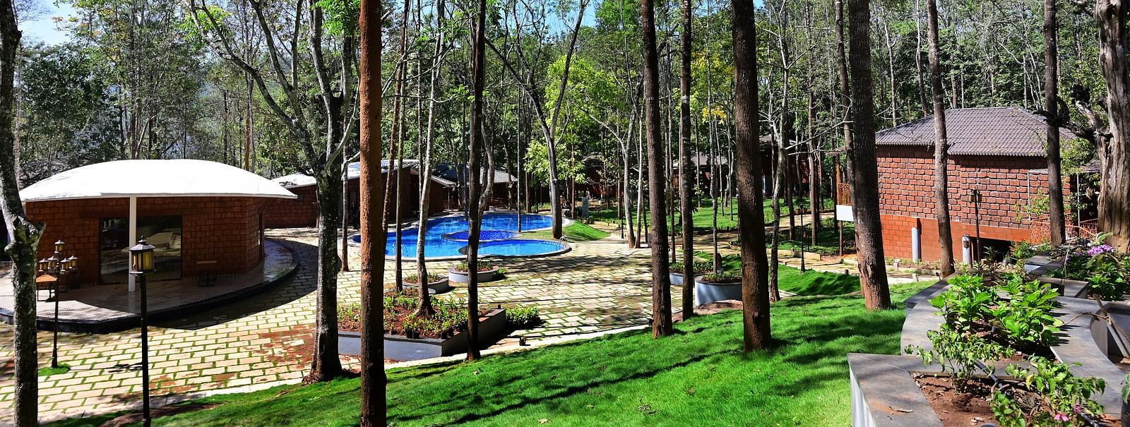 An outdoor area at TGI Redolent Resort, Kushalnagar, Coorg featuring trees, a pool, some cottages, and lamp shades