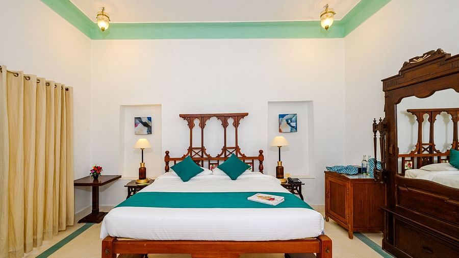 A bedroom with a wooden bed decorated with pillows and a bed runner, side lamps, framed art, and a wooden cabinet on the side. - Tijara Fort-Palace - 19th Century, Alwar