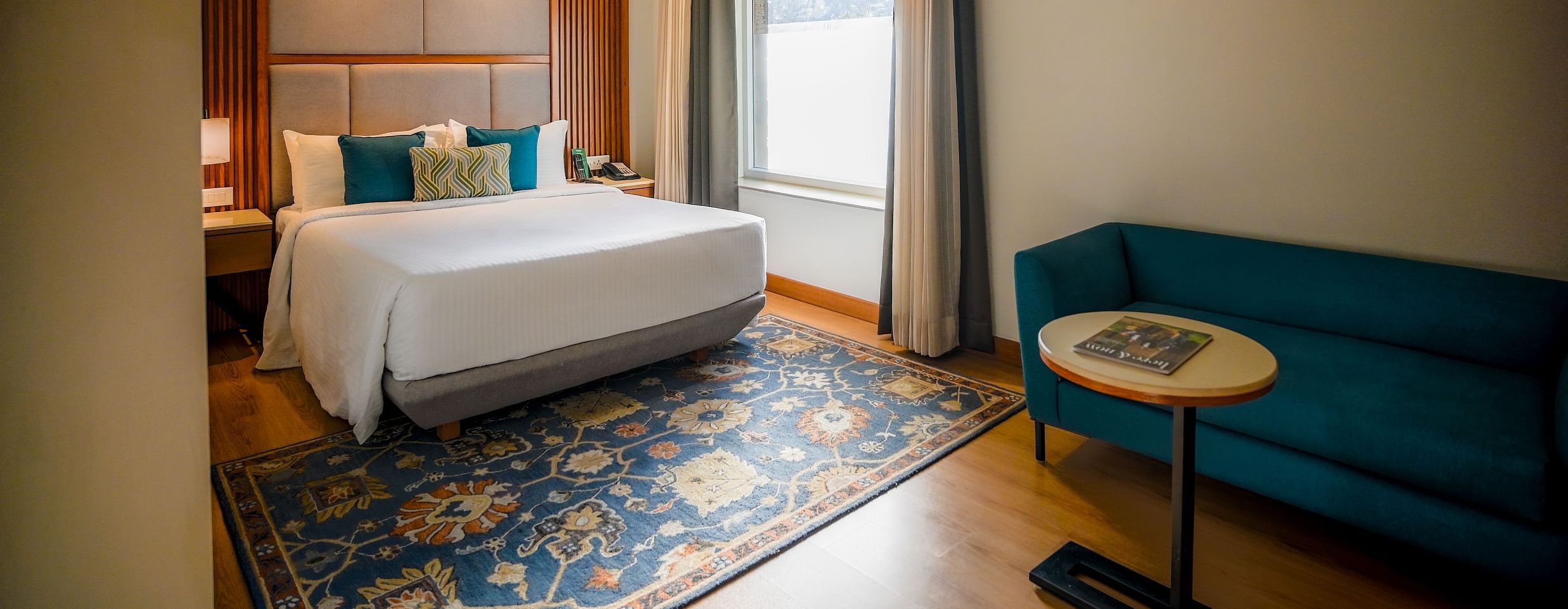 A brightly lit hotel room features a neatly made bed with white linens, a teal armchair, and a small round side table with a lamp. A window with sheer curtains allows natural light to filter in - Ananta Spa and Resort, Pushkar
