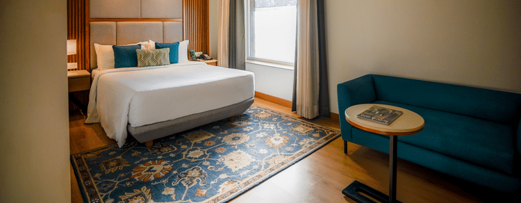 A brightly lit hotel room features a neatly made bed with white linens, a teal armchair, and a small round side table with a lamp. A window with sheer curtains allows natural light to filter in - Ananta Spa and Resort, Pushkar