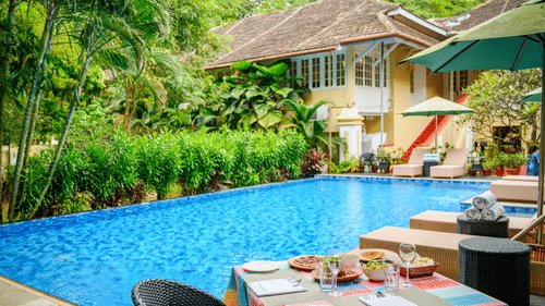 A dining table set by the swimming pool at The Tower House, surrounded by greenery, overlooking the buildings in the property