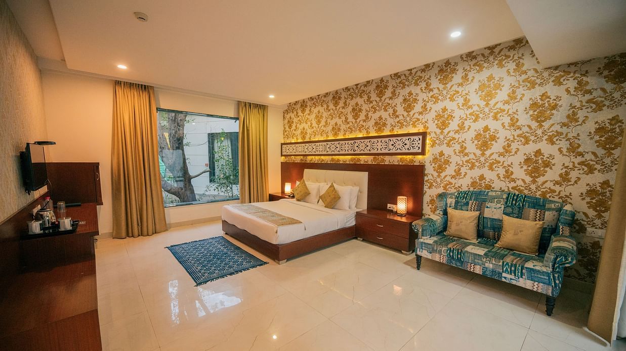 Wide view of a luxurious hotel room featuring a king-size bed, a stylish blue sofa, a dark wooden dresser, and large windows with sheer curtains - Clarks Safari, Ranthambore