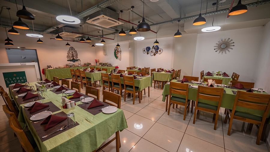 Clarks Safari Kheda's dining area with multiple tables are set with neat coverings and chairs, lights, neatly arranged tissues, plates, spoons, a small money plant on the centre, service counter, ac, and decorative elements on the walls.