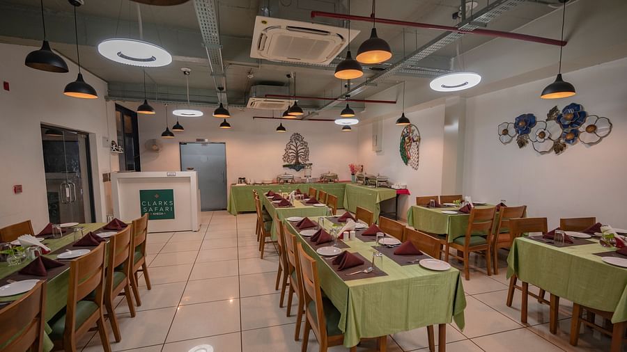 Clarks Safari Kheda's dining area with multiple tables are set with neat coverings and chairs, lights, neatly arranged tissues, plates, spoons, a small money plant on the centre, service counter, ac, and decorative elements on the walls.