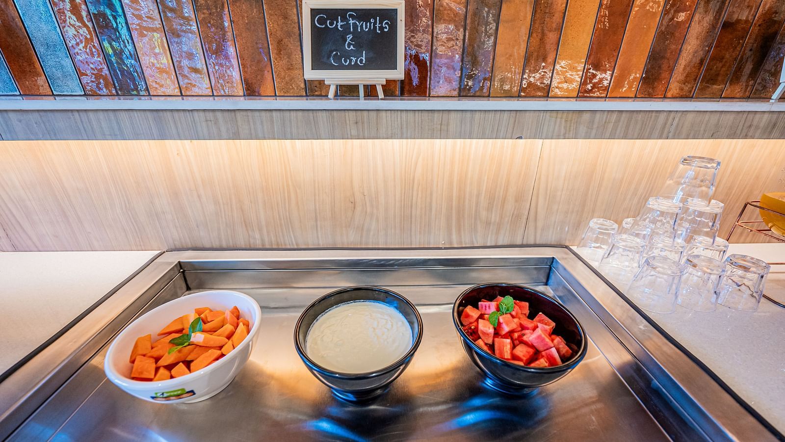 A counter at Indie Stays Mumbai International Airport, featuring three bowls filled with fresh vegetables and fruits on a tray, with glassware placed beside it.