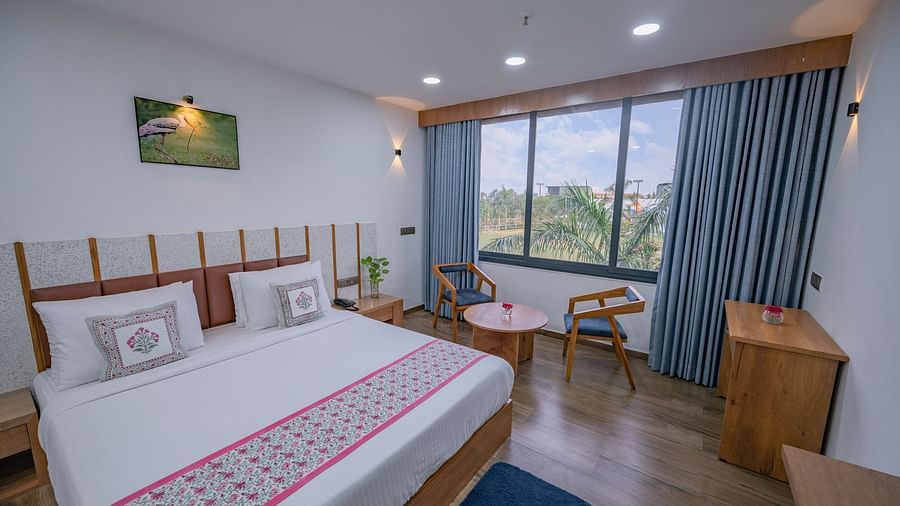 The view of a double bedroom at Clarks Safari Kheda with wooden flooring, frame, large window with curtains, tv, two chairs, round table, desk, remote, flower vase on top of the table and neatly arranged pillows.