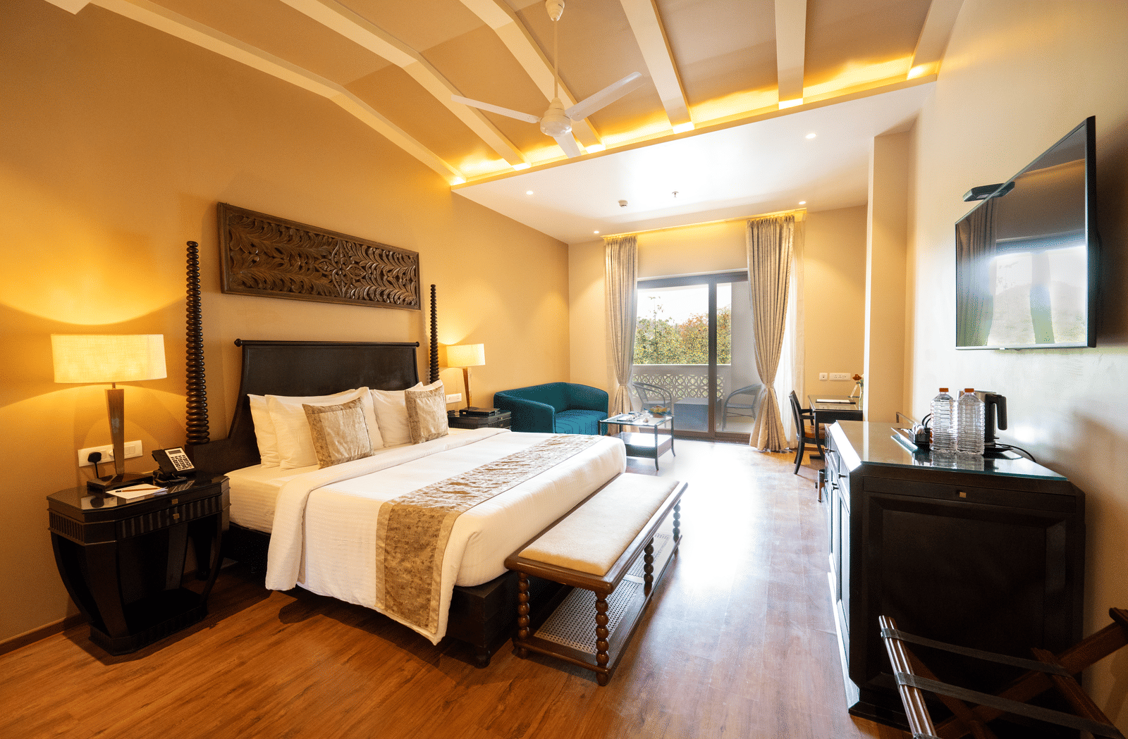 Premium Room at Essentia Luxury Resort & Spa, Udaipur showing a plush bed and an ottoman at its foot facing the TV over a table on the wall with a seating space by the door to the balcony