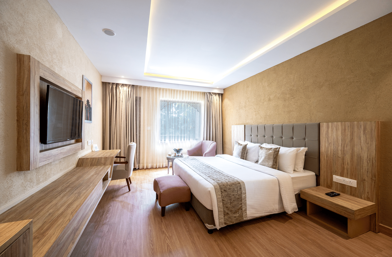 Standard Room at Essentia Luxury Resort & Spa, Udaipur featuring a plush bed witrh ottoman facing the TV on the wall over a running desk with a chair by the window with a seating area