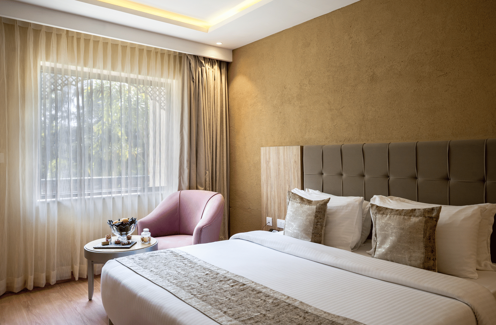 Standard Room at Essentia Luxury Resort & Spa, Udaipur featuring a plush bed with pillows, cushions and a bed runner next to a seating space by the window with sheer curtains